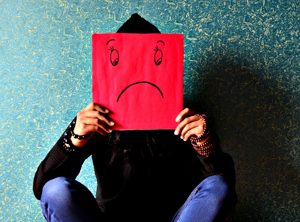 Woman Holds a Red Carboard Over Her Face With a Sad Smily Face Drawn in Black Marker